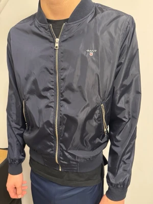 Mörkblå bomberjacka från GANT - Snygg mörkblå bomberjacka från GANT med glansig finish och ribbade muddar. Jackan har dragkedja framtill, två sidofickor med dragkedja och diskret GANT-logga på bröstet. Står att det är storlek M men detta är barnstorlek skulle säga det är 146/152