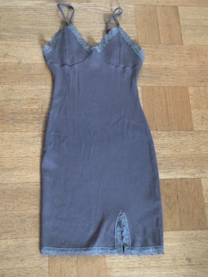 Mörkgrå kort klänning med spets - Kort, mörkgrå klänning med tunna axelband och v-ringad hals. Klänningen har spetsdetaljer vid urringningen, nederkanten och en liten slits framtill. Tillverkad i ribbat material för en slim och bekväm passform.