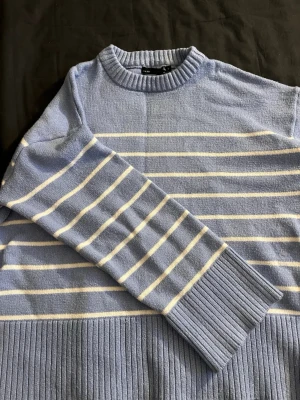 Blåvit randig stickad tröja - Mysig ljusblå stickad tröja med vita horisontella ränder. Tröjan har lång ärm, ribbade muddar och nederkant samt en klassisk rund halsringning. Perfekt för dig som gillar en avslappnad men stilren look.