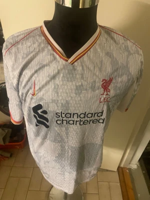 Liverpool bortatröja Nike M.Salah 11 - Snygg Liverpool FC bortatröja från Nike med M.Salah och nummer 11 på ryggen. Tröjan är ljusgrå med subtilt mönster, röda och gula detaljer vid krage och ärmslut samt klubbmärke och sponsortryck framtill. Perfekt för dig som älskar fotboll och vill stötta ditt lag. Den är size xxl men passar xl samt L