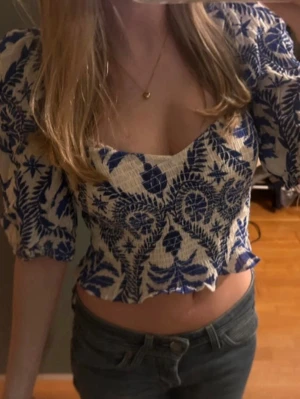 Blåvit blommig croppad blus - Trendig croppad blus med blått blommönster på vit botten. Puffiga korta ärmar och vågig nederkant ger en romantisk vibe. Tunn, luftig bomull som passar perfekt till jeans eller kjol. Snygg v-ringning framtill.