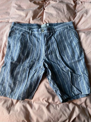Blå randiga shorts Dressmann - Snygga blå shorts från Dressmann med vita vertikala ränder. Klassisk passform med knapp och dragkedja framtill samt fickor på sidorna. Perfekta för varma dagar och en avslappnad stil. Små i storlek 