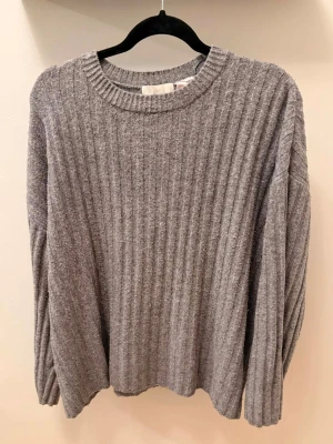 Grå ribbstickad tröja - Mysig grå stickad tröja med ribbad struktur och rund halsringning. Tröjan har långa ärmar och en avslappnad passform, perfekt för lager på lager. Materialet känns mjukt och bekvämt mot huden.