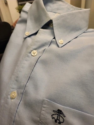 Ljusblå skjorta från Brooks Brothers  S - Klassisk ljusblå skjorta från Brooks Brothers med button-down krage och vit knappstängning. Skjortan har en bröstficka med mörkblå broderad logga och är tillverkad i bomull. Perfekt för en clean och stilren look.