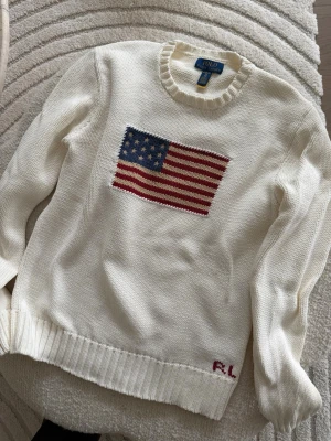 Vit stickad tröja Polo Ralph Lauren storlek 170 (XL) - Vit stickad tröja från Polo Ralph Lauren med amerikansk flagga i blått, rött och vitt på bröstet samt RL-broderi nertill. Tröjan har rund halsringning och ribbade muddar vid ärmslut och nederkant. Perfekt för dig som gillar klassisk preppy stil. Finns några små fläckar se bilder, går säkert att tvätta bort. 