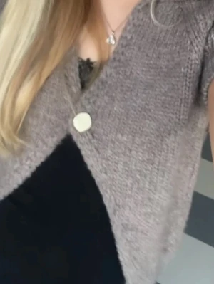 Kortärmad stickad Zara kofta i gråbeige - Kort, stickad kofta i gråbeige nyans med en stor dekorativ knapp framtill. Koftan har V-ringning och är gjord i ett mjukt, fluffigt material. Perfekt att slänga över en topp för en chill look.