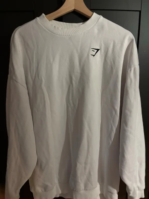 Vit Gymshark sweatshirt - Vit sweatshirt från Gymshark med rund halsringning och svart logga på bröstet. Tröjan har ribbade muddar vid ärmslut och nederkant och är gjord i mjukt bomullsmaterial. Perfekt för träning eller chill.