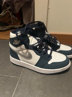 Air Jordan 1 Mid blå/vit/silver - Säljer ett par Air Jordan 1 Mid sneakers i blått, vitt och silver. Skorna har klassisk hög siluett, blå snörning och detaljer i silver på swoosh-loggan. Ovandelen är i läder med perforerad tåbox för extra ventilation. Perfekta för dig som gillar streetwear och snygga sneakers.