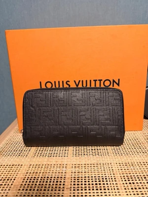 Svart plånbok från Fendi med mönster - Snygg svart plånbok från Fendi med präglat FF-mönster över hela ytan. Plånboken har dragkedja runtom och en dekorativ detalj med Fendi-logga på baksidan. Tillverkad i läder och har en stilren, rektangulär form som rymmer det mesta du behöver. Ena dragkedjan så har den man öppnar med försvunnit, men det finns med en läder strap man kan lägga på.