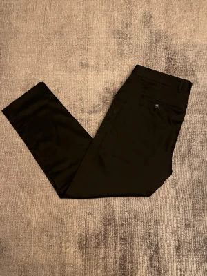 Svarta kostymbyxor med rak passform - Snygga svarta kostymbyxor med rak passform och klassiska detaljer som bälteshällor, sidofickor och en bakficka med knapp. Materialet ser ut att vara slätt och något glansigt, perfekt för en stilren look. Passar till skjorta eller t-shirt för en clean outfit.