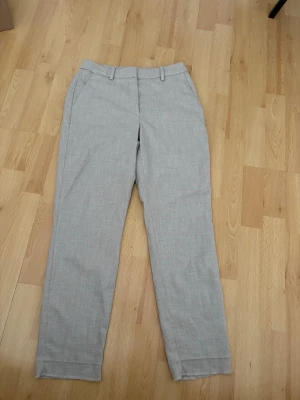 Ljusgrå kostymbyxor med rak passform - Snygga ljusgrå/beige  kostymbyxor med rak passform och pressveck. Byxorna har hällor för bälte och sidfickor. Perfekta för en clean och stilren look. Materialet känns lätt och passar bra till olika outfits.de är gansks slimfit så som slacks.