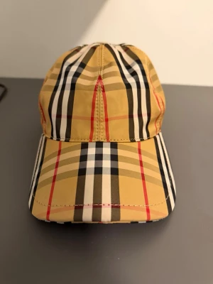 Burberry keps - Säljer en ikonisk keps från Burberry med det klassiska beige, svarta, vita och röda rutmönstret. Kepsen har böjd skärm och justerbart spänne bak i svart. Tillverkad i bomullsmix som känns lätt och bekväm på huvudet. Perfekt accessoar för en trendig streetwear-look.