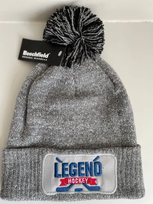 Grå Legend Hockey mössa med tofs - Säljer en grå stickad mössa med svartvit tofs på toppen. Perfekt för dig som gillar sportig stil. En lite snyggare broderad patch framtill. Från beechfield.