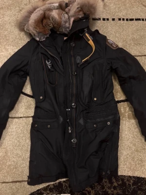 Svart Parajumpers parkas med päls - Säljer en svart parkas från Parajumpers med stor luva kantad av äkta päls. Jackan har flera fickor, gul detaljrem vid halsen och klassisk PJS-patch på ärmen. Perfekt för kalla dagar och riktigt snygg streetstil.