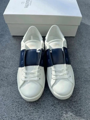 Valentino Garavani vita sneakers - Snygga vita sneakers från Valentino Garavani med bred mörkblå läderdetalj över vristen. Klassisk låg modell med vita snören och diskret logga på plösen. Tillverkade i slätt läder för en clean och exklusiv look.