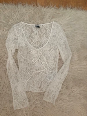 Vit spetsblus med snörning - Säljer en superfin vit spetsblus från Gina med lång ärm och snygg snörning framtill. Blusen är genombruten med blommigt spetsmönster och har en v-ringad urringning. Perfekt för dig som vill ha en trendig och feminin topp.
