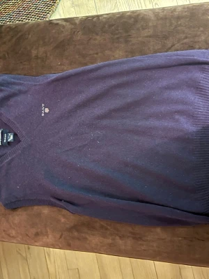  stickad tröja från GANT - Mysig lila stickad tröja från GANT med v-ringning och ribbade muddar. Tröjan har en liten broderad logga på bröstet och är tillverkad i mjuk lammull. Perfekt för dig som gillar klassisk och stilren design.