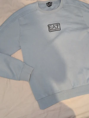Ljusblå EA7 tröja - Säljer en stilren ljusblå sweatshirt från EA7 Emporio Armani. Tröjan har rund halsringning, ribbade muddar och en snygg logga på bröstet. Perfekt för dig som gillar enkel men trendig streetwear.