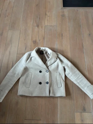 Beige kort rock från ONLY - Stilren beige kort rock från ONLY med dubbelknäppning och två framfickor. Klassisk krage och raka ärmar. Perfekt för dig som gillar en clean och modern look. Materialet känns mjukt och har en ull-liknande finish.