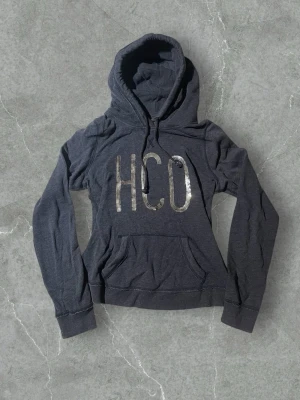 Mörkgrå hoodie från HCO Hollister | 36/38 - Kolla måtten (finns i bilderna)! Mörkgrå hoodie från Hollister med stor huva och magficka. Framsidan har stora, glittriga bokstäver 'HCO' i silver. Tillverkad i mjukt bomullsmaterial och har långa ärmar med ribbade muddar. Perfekt för en avslappnad och trendig stil.