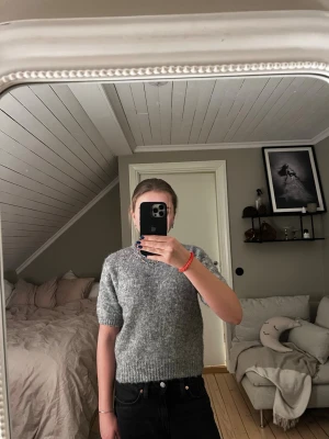 Grå stickad t-shirttröja - Mysig grå stickad t-shirttröja med korta ärmar och rund hals. Tröjan har en rak passform och ribbstickade muddar vid ärmslut. Enkel att styla till jeans eller kjol.