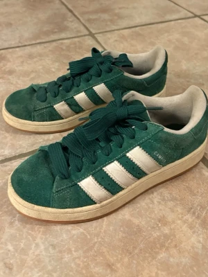 Gröna Adidas Campus sneakers - Säljer ett par gröna Adidas Campus sneakers i mocka med klassiska vita ränder på sidorna och vit sula. Skorna har gröna skosnören och låg profil. Perfekta för dig som gillar retrostil och streetwear.