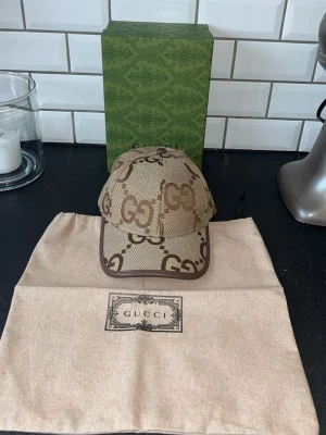 Beige keps med GG-logga från Gucci - Säljer en beige keps från Gucci med klassiskt GG-monogram i brunt över hela kepsen. Kepsen har böjd skärm, bruna kanter och är tillverkad i canvasmaterial. Perfekt för dig som vill ha en stilren accessoar med lyxig känsla.