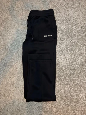 Svarta mjukisbyxor från Les Deux - Snygga svarta mjukisbyxor från Les Deux med raka ben och dragsko i midjan. Byxorna har diskreta fickor och en liten vit logga på vänster sida. Perfekta för en chill och stilren look. Materialet känns mjukt och bekvämt.