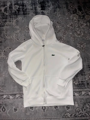 Vit zip hoodie från Lacoste - Snygg vit hoodie från Lacoste med dragkedja framtill och klassisk grön krokodil-logga på bröstet. Hoodien har huva, ribbade muddar och fickor framtill. Perfekt för en clean och stilren look. Materialet känns mjukt och bekvämt.