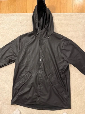 Svart regnjacka med huva - Stilren svart regnjacka med huva och dragsko, knäppning med tryckknappar och två snedställda fickor framtill. Jackan har en rak passform och är tillverkad i vattenavvisande material, perfekt för regniga dagar.