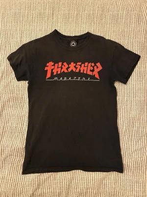Svart Thrasher t-shirt med tryck - Säljer en svart vintage t-shirt från Thrasher med ett rött tryck på bröstet i japansk stil. Den är lite sliten med några hål, men den är vintage. Jag är 174 och den sitter bra på mig.