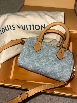 Louis Vuitton denim handväska - Nano Speedy - Säljer en snygg minihandväska från Louis Vuitton i ljusblå denim med klassiskt monogrammönster. Väskan har beige läderdetaljer, guldfärgade metalldelar och avtagbar axelrem. Perfekt för dig som gillar trendiga accessoarer med lyxig känsla.