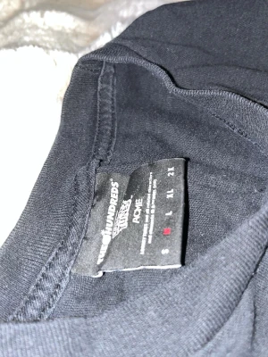 Svart T-shirt The Hundreds x Looney Tunes - Säljer en svart T-shirt från The Hundreds i samarbete med Looney Tunes och ACME. T-shirten har en klassisk passform, rund hals och är tillverkad i mjuk bomull. På ryggen finns en vit ACME-bombgrafik som ger en unik streetwear-vibe.