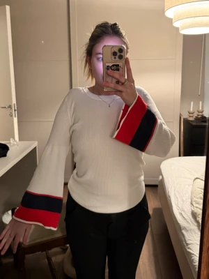 Vit tröja  tommy hilfiger - Vit ribbstickad tröja med vida ärmar och breda ränder i rött och mörkblått längst ner på ärmarna. Tröjan har en rund halsringning och är tillverkad i ett mjukt stickat material. Perfekt för dig som gillar unika detaljer och färgkombinationer. Tommy hilfiger 
