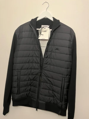 Svart cardigan från J.Lindeberg - Helt ny och oanvänd cardigan från j Lindberg kvitto och lappar finns. Snygg svart hybridjacka från J.Lindeberg med quiltad front och stickade ärmar. Jackan har dragkedja framtill, två sidofickor med dragkedja och diskret logga på bröstet. Perfekt för lager-på-lager och stilren look.