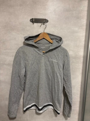 Grå hoodie från Calvin Klein - Snygg grå hoodie från Calvin Klein med vit logga på bröstet. Hoodien har långa ärmar, huva och en vit samt svart rand längst ner vid fållen. Tillverkad i mjukt och tunt material som känns skönt mot huden.