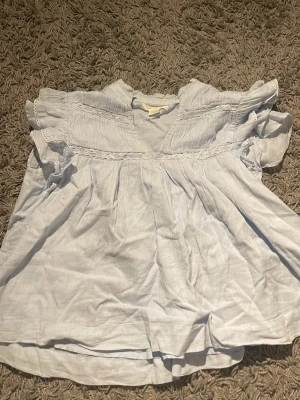 Ljusblå volangblus från Gina Tricot - Supersöt ljusblå blus från Gina Tricot med volangdetaljer vid axlarna och v-ringning. Blusen har korta ärmar och är gjord i ett tunt, luftigt tyg.