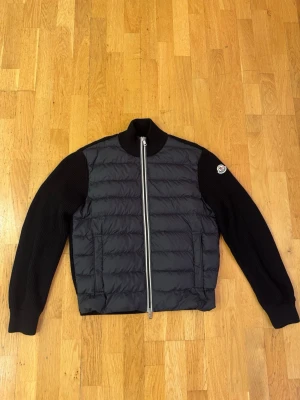 Svart cardiganjacka från Moncler - Snygg svart cardiganjacka från Moncler med quiltad front och stickade ärmar. Jackan har hög krage, dragkedja framtill och två fickor. Moncler-logga på vänster ärm. Perfekt för lager-på-lager och streetwear-stil.