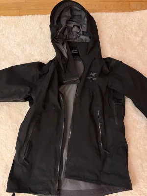 Arc'teryx Beta Ar jacket W black M - Snygg svart regnjacka från Arc'teryx med minimalistisk design och justerbar huva. Jackan är tillverkad i slitstarkt GORE-TEX-PRO-material som står emot regn och vind. Perfekt för utomhusaktiviteter och äventyr. Dragkedjor framtill och vid fickor.