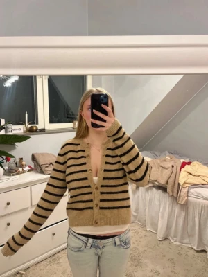 Beige och svart randig kofta - Mysig beige kofta med svarta horisontella ränder och v-ringning. Koftan har knappar framtill, ribbade muddar och är något croppad i modellen. Perfekt att slänga över ett linne eller t-shirt för en avslappnad look.