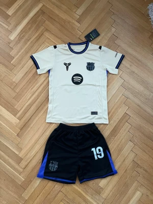 FC Barcelona Lamine Yamal matchställ - Snyggt FC Barcelona matchställ med Lamine Yamal #19. Tröjan är beige med svarta och blå detaljer, klubbmärke och sponsorloggor framtill. Shortsen är svarta med blå inslag och nummer 19. Perfekt för fotbollsträning eller match.