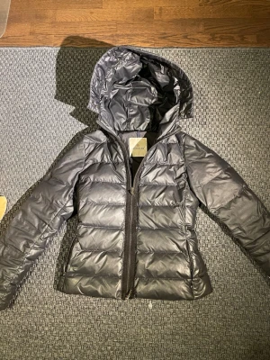 Svart dunjacka från Moncler med huva - Snygg svart dunjacka från Moncler med quiltad design och fast huva. Jackan har dragkedja framtill och två sidofickor. Perfekt för kalla dagar och ger en stilren look. Klassisk Moncler-logga på insidan. Passar dig som vill ha både värme och stil.