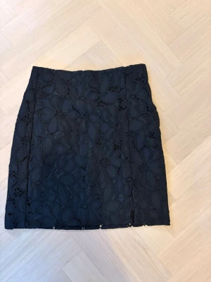 Svart spetskjol med blommönster - Kort svart kjol med blommig spets och rak passform. Kjolen har ett elegant och stilrent utseende med tydlig struktur i spetsen. Perfekt för dig som vill ha en enkel men ändå detaljrik kjol.