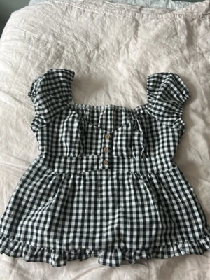 Rutig svartvit topp! - Supersöt blus i svartvitt rutigt mönster med korta puffärmar, knappar framtill och volangkant! Perfekt till sommaren! Passar allt från S-M
