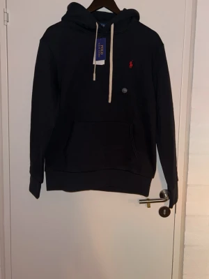 Svart hoodie från Polo Ralph Lauren - Snygg svart hoodie från Polo Ralph Lauren med klassisk röd logga broderad på bröstet. Hoodien har huva med vita dragsnören och en stor magficka framtill. Tillverkad i mjukt bomullsmaterial som känns skönt mot huden. Perfekt för en avslappnad och stilren look. Hejsan alla! Jag har precis fyllt 18 år och har fått en massa presenter. Det är mycket av presenterna som inte passar min stil, men det är väldigt fina och de som bär dessa kläderna har fin stil. Det är därför jag väljer att sälja detta. 