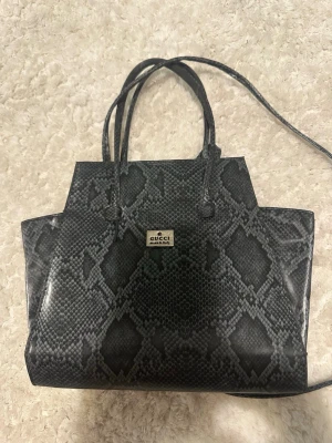Gucci handväska i ormmönstrat läder - Snygg handväska från Gucci i svart och grått ormmönstrat läder. Väskan har två smala handtag och en längre axelrem för olika bärstilar. Metallbricka med Gucci-logga framtill. Perfekt för dig som vill ha en statementväska med lyxig känsla.