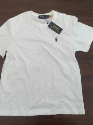 Vit t-shirt från Polo Ralph Lauren - Vit klassisk T-shirt från Polo Ralph Lauren i mjuk och skön bomull. Stilren modell med rund hals och den ikoniska broderade logotypen på bröstet. Perfekt basplagg som passar till allt – både vardag och mer uppklätt.  🆕 Ny med prislapp kvar ⚠️ Litet fläck på baksidan, går troligen bort i tvätten 🚚 Snabb leverans 💬 Tar bud