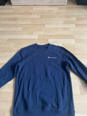Mörkblå sweatshirt från Champion - Mörkblå sweatshirt från Champion med klassisk logga broderad på bröstet. Tröjan har rund halsringning, långa ärmar och ribbade muddar vid ärmslut och nederkant. Perfekt för en avslappnad och sportig stil.