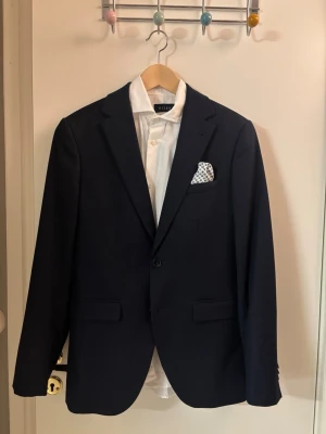 Mörkblå kostym från Raily  - Snygg mörkblå kostym från Brothers i märket Riley med klassisk kavaj och matchande byxor. Kavajen har två knappar, fickor och en vit näsduk med prickigt mönster i bröstfickan. Perfekt för dig som vill ha en stilren och tidlös look.