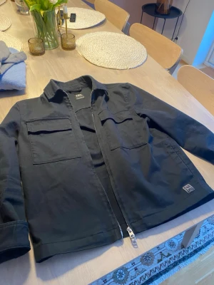 Svart overshirt från Zara - Stilren svart overshirt från Zara med två stora bröstfickor och dragkedja framtill. Overshirten har klassisk krage och är tillverkad i ett kraftigare bomullstyg som ger en avslappnad men snygg look. Perfekt att slänga över en t-shirt.